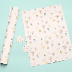 Op de afbeelding: Een rol bloemen cadeaupapier en een plat vel bijpassend papier, plus twee ronde stickers, worden getoond op een lichtblauwe ondergrond. Het papier heeft een herhalend patroon van aquarelbloemen, cactussen en vazen in zachte pastelkleuren.