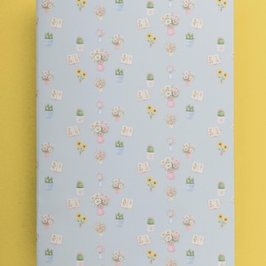 Op de afbeelding: Een lichtblauw notitieboek met een herhalend patroon van aquarel illustraties. Het patroon bevat roze vazen met bloemen, cactus in potten, zonnebloemen en open boeken. Het notitieboek is rechthoekig en heeft een matte afwerking.