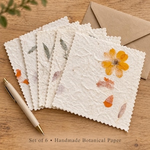 May include: A set of square, off-white handmade paper cards with deckled edges, featuring pressed yellow flowers and green leaves embedded in the texture. A light-coloured pen rests beside the cards, and a brown envelope is tucked behind the stack.