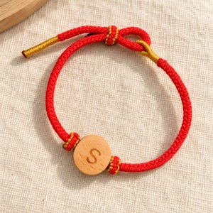Puede incluir: Una pulsera de cordón rojo con una cuenta de madera clara de forma circular grabada con la letra S. La pulsera se asegura con un mecanismo de nudo corredizo e incluye detalles de hilo metálico dorado en los extremos y alrededor de los nudos.