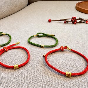 Könnte beinhalten: Vier verstellbare Kordelarmbänder in Rot und Grün, jedes mit einer goldenen zylindrischen Perle. Zwei Armbänder sind rot mit den Buchstaben A und E, zwei sind grün mit dem Buchstaben M und einem Symbol. Ein dekorativer Zweig mit roten Beeren liegt daneben.
