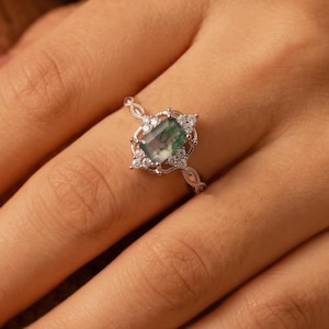Pode incluir: Um anel de tom prateado com uma pedra preciosa retangular verde-musgo no centro, rodeada por pequenas pedras transparentes de corte redondo. A banda apresenta um design metálico delicado e complexo que se estende a partir do centro.