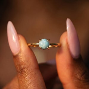 Könnte beinhalten: Ein goldener Ring mit einem runden, hellblauen Edelstein. Der Ring hat ein zartes Band und eine Vier-Zacken-Fassung. Der Edelstein ist der Mittelpunkt und zeigt seine hellblaue Farbe. Der Ring wird vor einem dunklen Hintergrund gehalten.