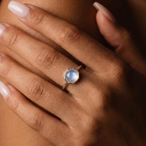 May include: A silver ring with a round, iridescent moonstone. The ring is set on a delicate band. The moonstone has a soft, glowing appearance. The ring is shown on a hand with manicured nails.