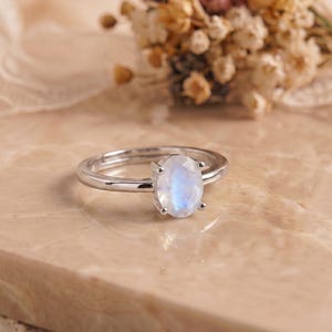 Peut inclure: Une bague argentée ornée d'un cabochon de pierre de lune de forme ovale, fixé par quatre griffes. L'anneau est lisse et poli, mettant en valeur un éclat irisé bleu subtil à l'intérieur de la pierre translucide, centrée sur la monture.