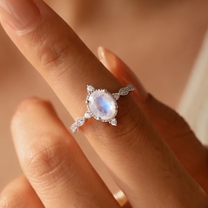 May include: A silver-toned ring featuring a central oval-shaped iridescent moonstone cabochon. The band is adorned with small, clear, round-cut stones set in a delicate, symmetrical pattern extending along the sides of the central setting.