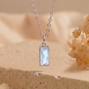 May include: A silver-toned necklace featuring a rectangular pendant with a faceted, iridescent light blue stone. The pendant hangs from a delicate silver-toned chain with circular links, showcasing a clear, reflective surface and metallic frame.