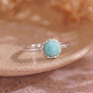 May include: A silver ring with a round, light blue gemstone. The gemstone is set in a decorative bezel. The ring is displayed on a neutral-toned surface, with a soft, blurred background.