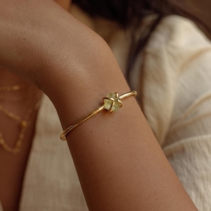 Könnte beinhalten: Ein goldfarbenes Armreif-Armband mit einem hellgrünen Edelstein in der Mitte. Das Armband wird am Handgelenk einer Person getragen, wobei der Fokus auf dem Schmuck liegt. Der Hintergrund ist unscharf, wodurch das Design und die Farbe des Armbands hervorgehoben werden.