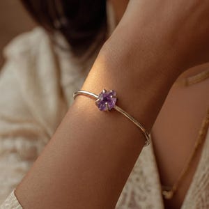 Può includere: Un bracciale rigido in metallo color argento con una singola pietra preziosa di ametista viola grezza fissata con griffe. Il bracciale ha una finitura liscia e lucida e un design circolare che si adatta saldamente al polso.