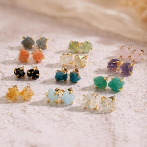 Puede incluir: Una colección de pendientes de botón con piedras preciosas en bruto y sin tallar en varios colores, incluyendo azul, verde, rosa, naranja, negro y transparente, cada una engastada en garras de metal de tono dorado.