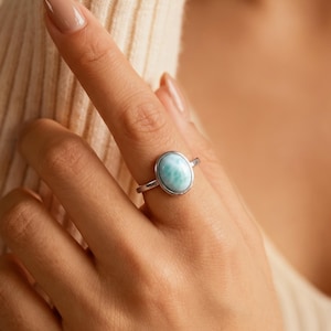 Könnte beinhalten: Ein Silberring mit einem glatten, ovalen, hellblauen Stein mit weißer Marmorierung. Der Stein ist in einer einfachen Metallfassung auf einem dünnen, polierten Silberband gefasst. Der Ring wird an einem Finger getragen und zeigt die Textur.
