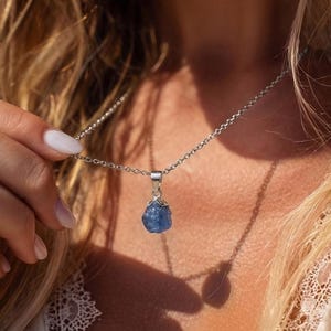 May include: A silver necklace with a small, rough-cut blue gemstone pendant. The pendant is suspended from a delicate chain. The necklace is worn against a background of fair skin and blonde hair.