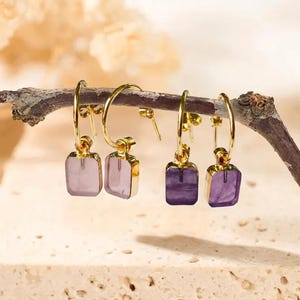 May include: Four pairs of gold-colored hoop earrings with square, gemstone pendants in shades of lavender and purple. The earrings are displayed on a small branch against a neutral background. The earrings are a jewelry item.