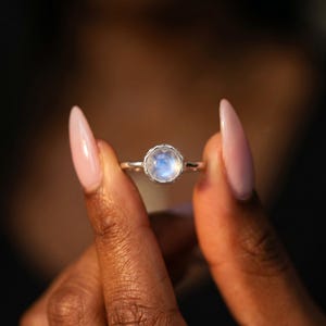 May include: A silver ring featuring a round, light blue moonstone. The gemstone is set in a textured bezel. The ring is held between two fingers with long, pink nails, against a blurred background.