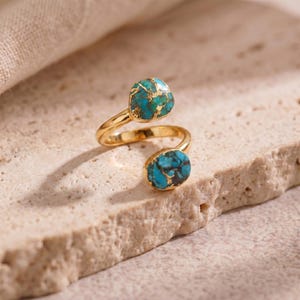 Pode incluir: Um anel aberto de tom dourado com duas pedras distintas de cor turquesa com veios dourados. O anel apresenta um design cruzado com uma pedra maior no topo e uma pedra menor na parte inferior, ligadas por uma banda fina e polida.