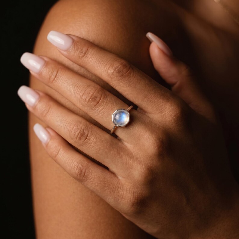 May include: A silver ring with a round, iridescent moonstone. The ring is set on a delicate band. The moonstone has a soft, glowing appearance. The ring is shown on a hand with manicured nails.