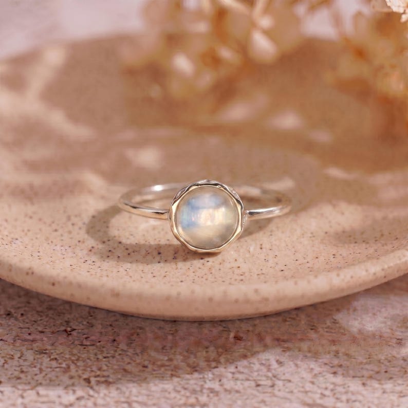 May include: A silver ring with a round, iridescent gemstone. The gemstone is set in a bezel with a scalloped edge. The ring is displayed on a light brown, textured surface, with soft, blurred background elements.
