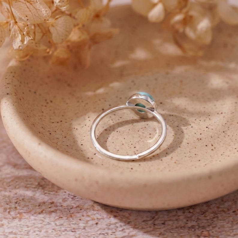 May include: A silver ring with a light blue gemstone. The ring is set on a light brown, speckled surface. The gemstone is oval-shaped and surrounded by a silver setting. The ring is a delicate piece of jewelry.