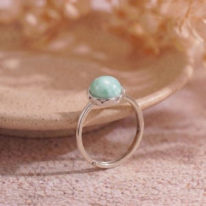 May include: A silver ring featuring a round, light green gemstone. The gemstone is set in a decorative bezel. The ring band is adjustable. The ring is displayed on a neutral background.