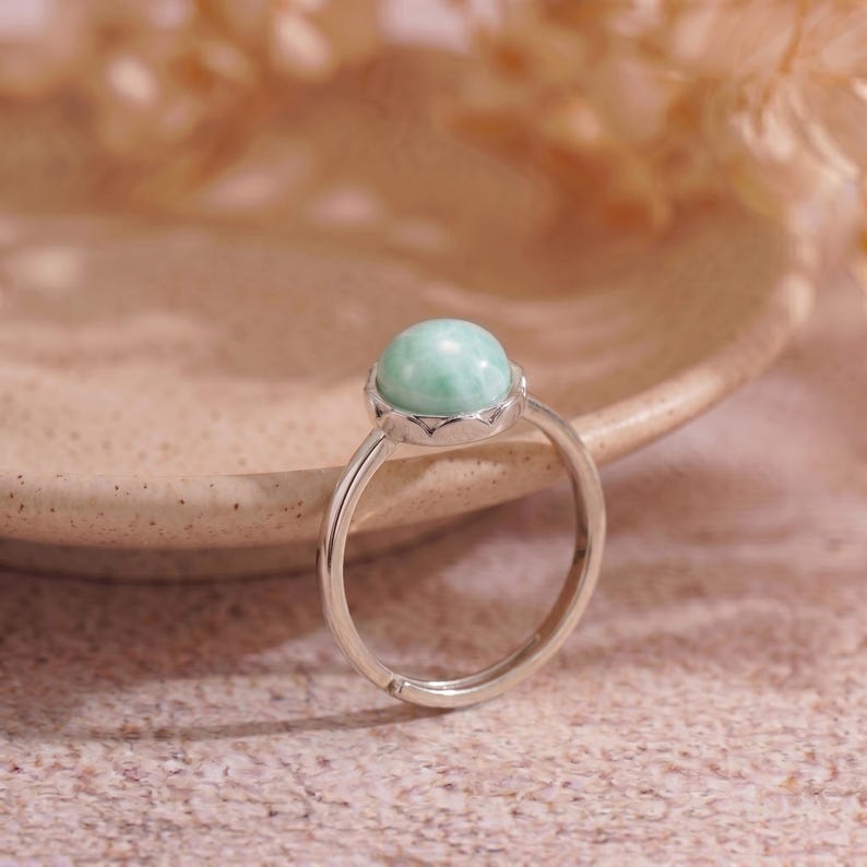 May include: A silver ring featuring a round, light green gemstone. The gemstone is set in a decorative bezel. The ring band is adjustable. The ring is displayed on a neutral background.