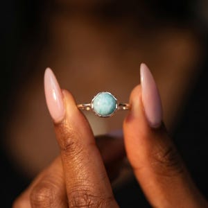 May include: A silver ring featuring a round, light blue gemstone. The gemstone is set in a scalloped bezel. The ring is held up by a person with long, pink fingernails.