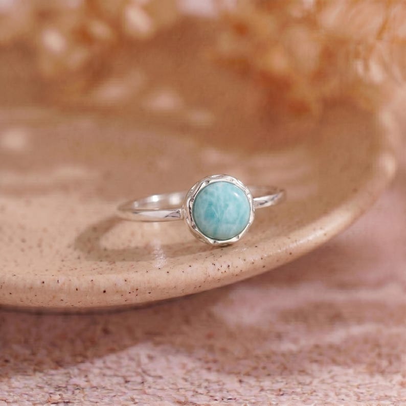 May include: A silver ring with a round, light blue gemstone. The gemstone is set in a decorative bezel. The ring is displayed on a neutral-toned surface, with a soft, blurred background.