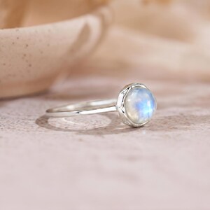 May include: A silver ring with a round, iridescent moonstone. The moonstone has a soft blue and white glow. The ring's band is thin and polished, and the stone is set in a decorative bezel.