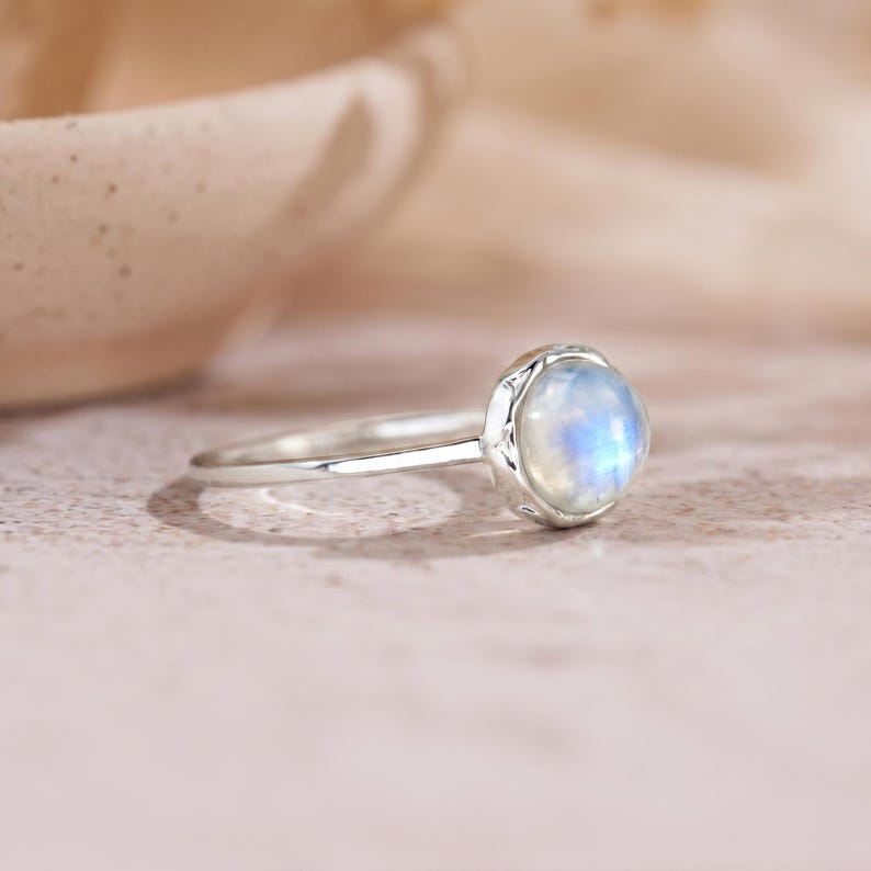 May include: A silver ring with a round, iridescent moonstone. The moonstone has a soft blue and white glow. The ring's band is thin and polished, and the stone is set in a decorative bezel.
