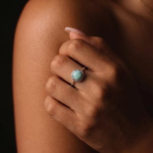 May include: A silver ring featuring a round, light blue gemstone. The ring is set in a delicate, ornate silver band. The gemstone has a soft, opaque appearance, and the ring is displayed on a hand.