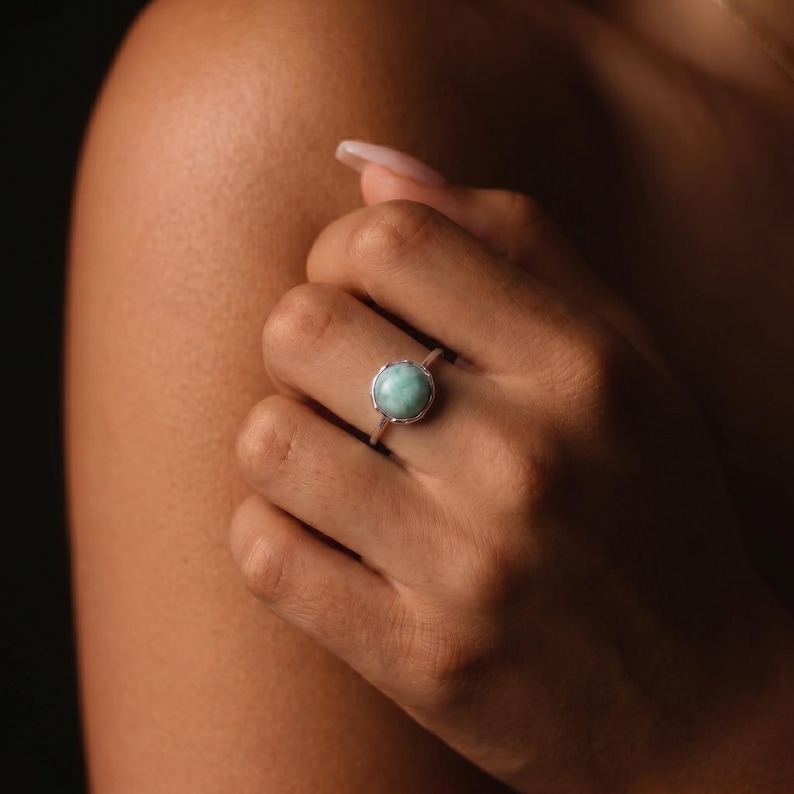 May include: A silver ring featuring a round, light blue gemstone. The ring is set in a delicate, ornate silver band. The gemstone has a soft, opaque appearance, and the ring is displayed on a hand.