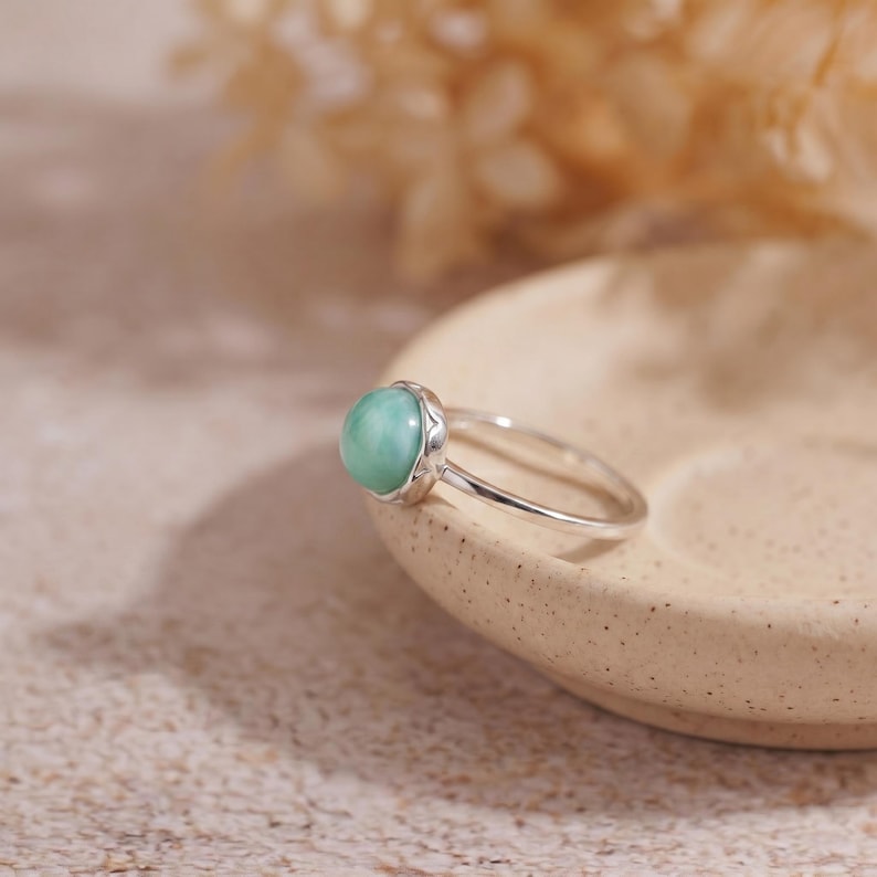 May include: A silver ring with a round, light green gemstone. The ring is set on a light-colored, speckled surface, with a blurred background. The gemstone is the focal point, showcasing a smooth, polished appearance. The ring's band is thin and delicate.