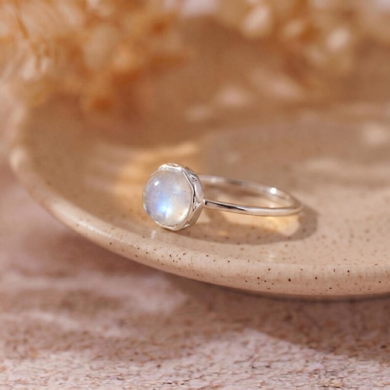 May include: A silver ring with a round, iridescent moonstone. The gemstone is set in a bezel setting, and the band is thin and polished. The ring is displayed on a neutral-toned surface, highlighting its elegant design.