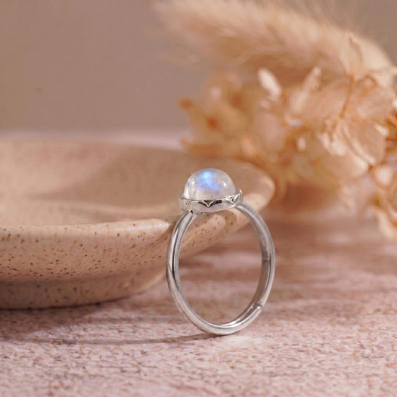 May include: A silver ring with a round, iridescent moonstone. The moonstone is set in a decorative bezel. The ring is displayed on a light pink surface, with a blurred background.