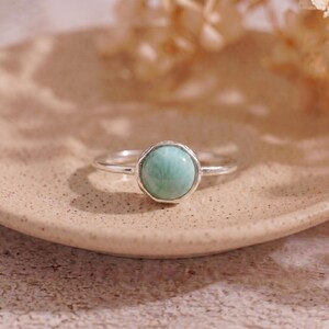 May include: A silver ring with a round, light green gemstone. The gemstone is set in a textured bezel. The ring is displayed on a beige plate, with soft, blurred floral elements in the background. The ring is a piece of jewelry.