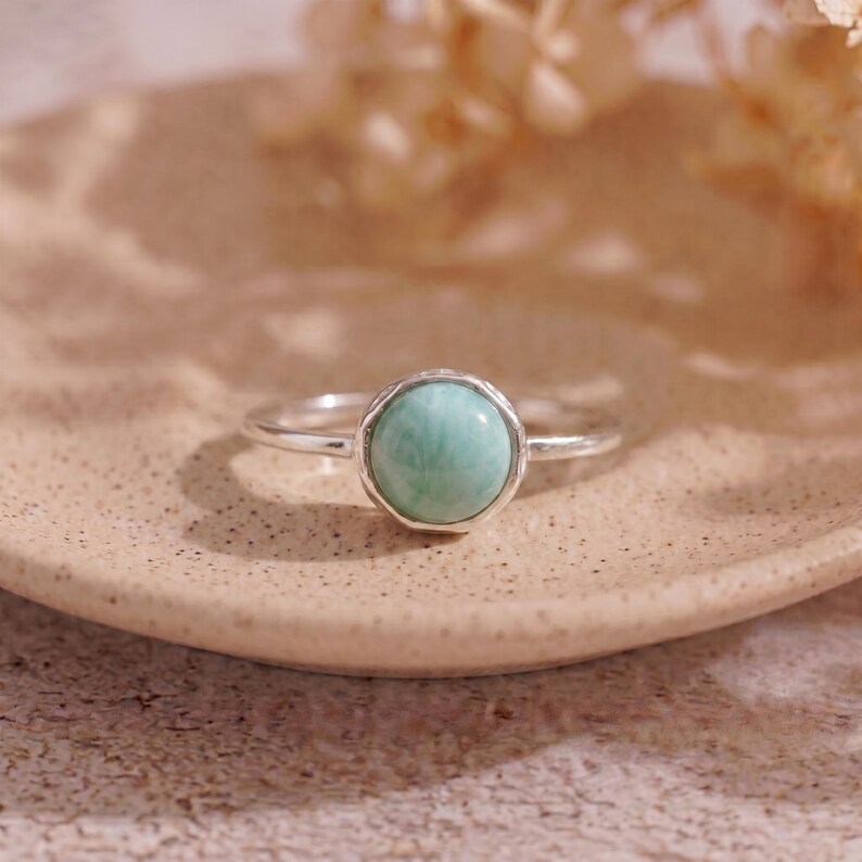 May include: A silver ring with a round, light green gemstone. The gemstone is set in a textured bezel. The ring is displayed on a beige plate, with soft, blurred floral elements in the background. The ring is a piece of jewelry.