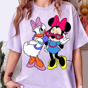 May include: A lilac t-shirt with a graphic of Disney's Daisy Duck and Minnie Mouse. Daisy Duck has a purple bow and Minnie Mouse wears pink sunglasses. The shirt has short sleeves.