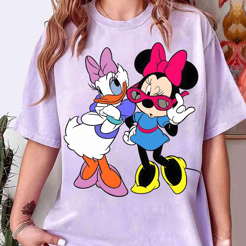 May include: A lilac t-shirt with a graphic of Disney's Daisy Duck and Minnie Mouse. Daisy Duck has a purple bow and Minnie Mouse wears pink sunglasses. The shirt has short sleeves.