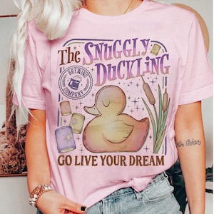 May include: Pink t-shirt with a graphic featuring a cartoon duck, the text "The Snuggly Duckling," and "Go Live Your Dream." The design includes a brewery logo, lanterns, and cattails. The shirt is a crew neck.