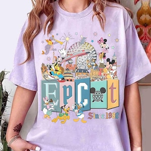 May include: A light purple t-shirt featuring a colorful graphic of Disney characters and landmarks. The word "EPCOT" is displayed in large, block letters, with "Since 1982" below. Mickey Mouse, Minnie Mouse, Donald Duck, and Goofy are among the characters.