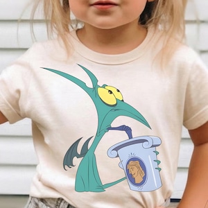 Puede incluir: Camiseta color crema con un personaje de dibujos animados verde azulado que sostiene una taza azul con un diseño de Hércules. El personaje tiene ojos amarillos grandes y un pico largo. La camiseta es una prenda infantil.