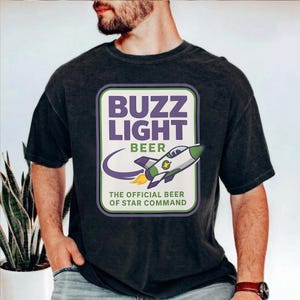 Puede incluir: Camiseta gris oscuro con un diseño rectangular que muestra el texto "Buzz Light Beer" y un cohete. El cohete es blanco y verde con llamas amarillas. El texto "The Official Beer of Star Command" está debajo.