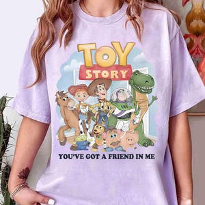May include: A light purple t-shirt featuring a colorful graphic of characters from the animated film "Toy Story." The design includes the words "TOY STORY" and "YOU'VE GOT A FRIEND IN ME."