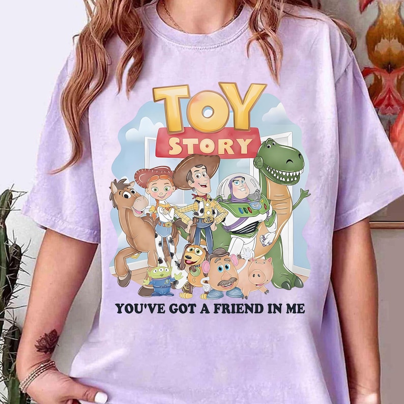 May include: A light purple t-shirt featuring a colorful graphic of characters from the animated film "Toy Story." The design includes the words "TOY STORY" and "YOU'VE GOT A FRIEND IN ME."