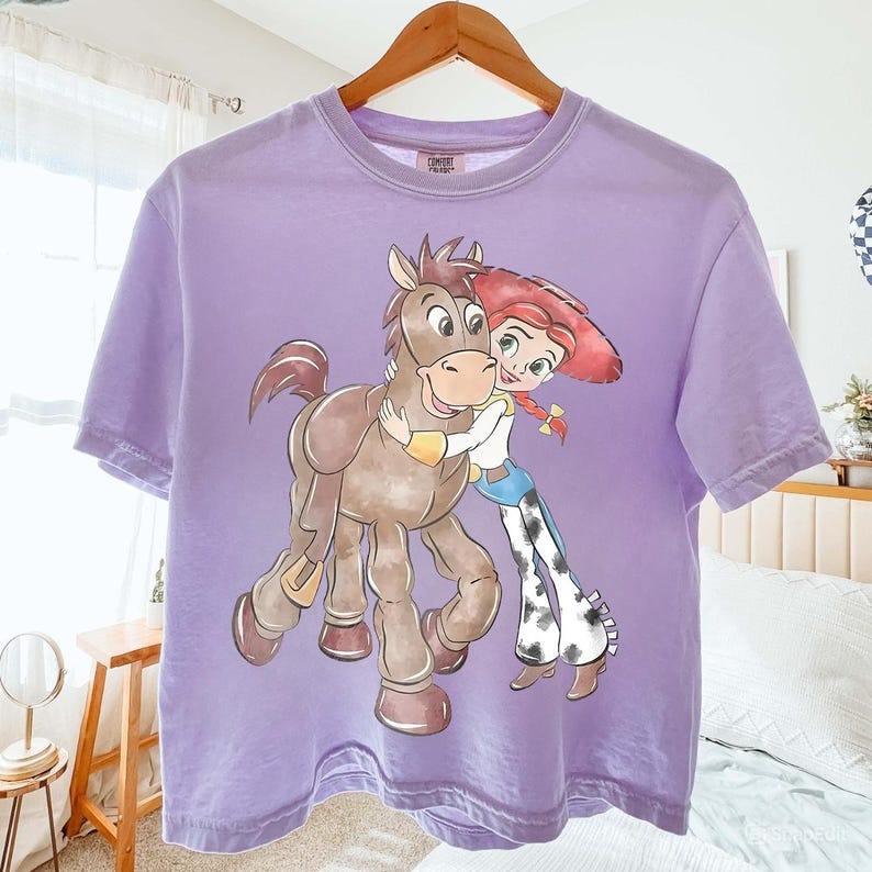 May include: A lavender t-shirt featuring a graphic of a cartoon horse and a cowgirl hugging. The cowgirl wears a red hat, blue jeans, and cow-print pants. The horse is brown with a yellow arm band.