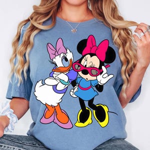 May include: A light blue t-shirt with a graphic of Daisy Duck and Minnie Mouse. Daisy Duck has a purple bow, and Minnie Mouse wears a pink bow and sunglasses. The cartoon characters are in a colourful design.