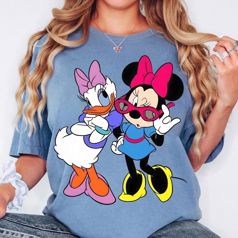 May include: A light blue t-shirt with a graphic of Daisy Duck and Minnie Mouse. Daisy Duck has a purple bow, and Minnie Mouse wears a pink bow and sunglasses. The cartoon characters are in a colourful design.