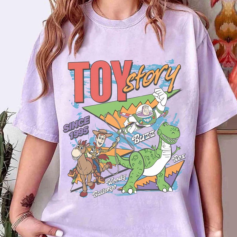 May include: A light purple t-shirt featuring a colorful graphic of characters from the animated film "Toy Story." The design includes Woody, Buzz Lightyear, Rex the dinosaur, and the text "Toy Story" and "Since 1995."