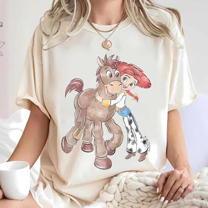 May include: Cream-colored t-shirt featuring a cartoon illustration of a character hugging a brown horse. The character has red hair and is wearing a cowboy hat, blue jeans, and a white shirt. The horse is brown with a yellow harness.