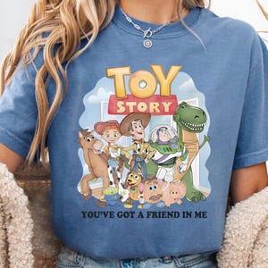 May include: Blue t-shirt featuring a colorful graphic of characters from the animated film "Toy Story." The design includes the words "TOY STORY" and "YOU'VE GOT A FRIEND IN ME."
